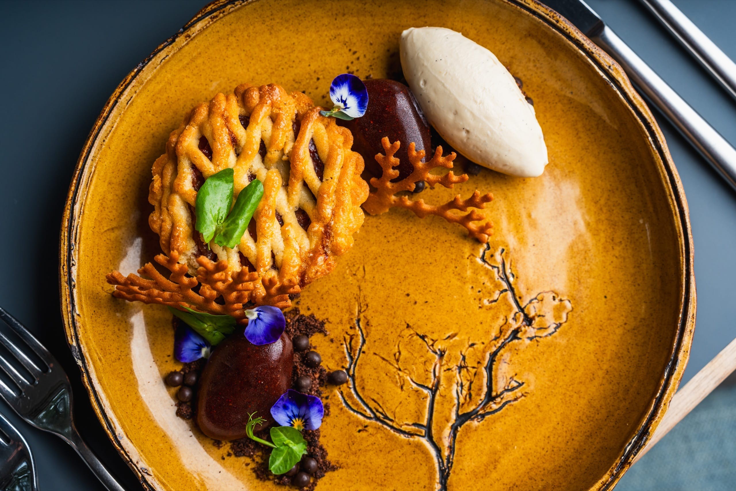 A lace-tuile dessert with poached fruit, an ice-cream quenelle and edible flowers on an amber plate, viewed from above